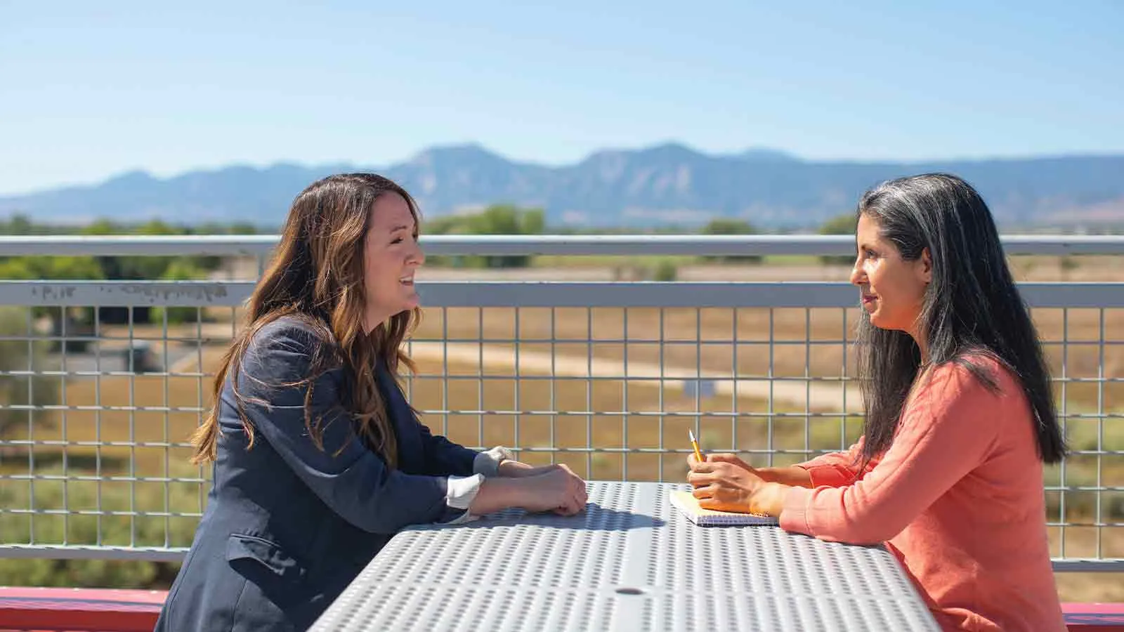 Two women seated across an outdoor table with mountains on the horizon, one taking notes, the kind of client conversation that follows a report showing the ownership form actually of record