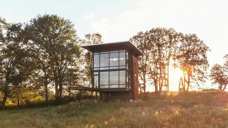 A narrow two-storey glass and weathered-steel house standing alone on a grassy hillside at sunset, the kind of isolated collateral a lender may end up taking back by deed