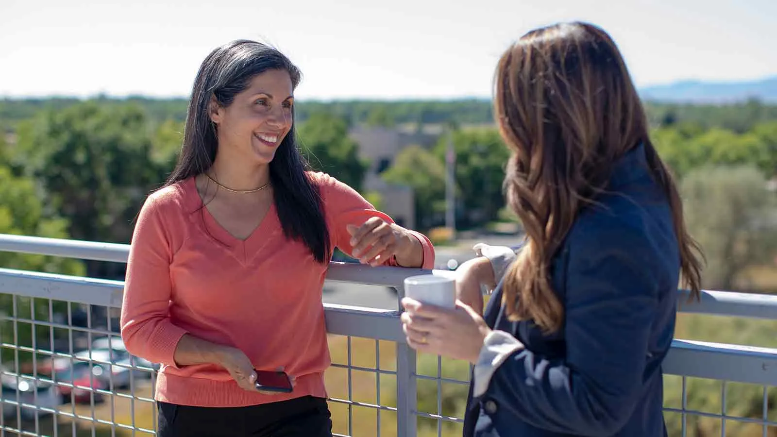 Two women talking over a balcony railing with trees and rooftops behind them, the kind of conversation where a borrower and a lender decide whether to convey rather than foreclose